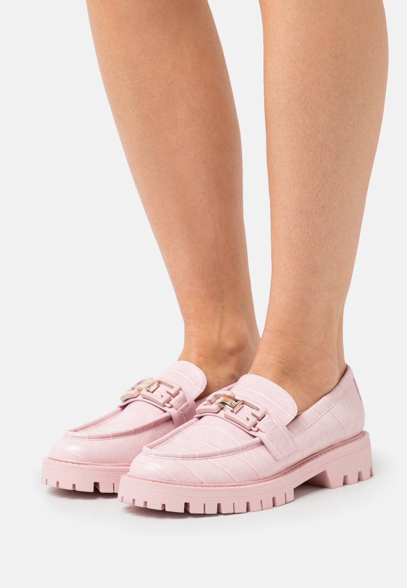 Mocassins roses à semelles épaisses avec une texture embossée croco et un détail de boucle en métal portés pieds nus, vus de côté et de face.