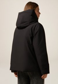 Zwarte waterdichte jas met een grote capuchon, met een relaxte pasvorm en horizontale naaddetails. Het materiaal lijkt glad en duurzaam.