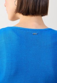 Personne aux cheveux courts bruns portant un haut bleu vif vue de dos, montrant une petite étiquette métallique près de l'encolure.