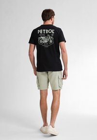 Črna bombažna majica s teto grafiko motorja in besedilom "PETROL" na hrbtni strani. V kombinaciji s svetlo zelenimi kratkimi hlačami. Priložnostni stil.