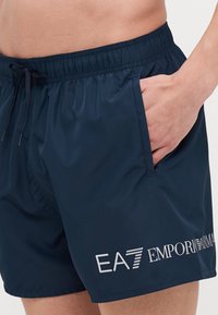 Kék úszónadrág sima anyagból, rugalmas derékpánttal, húzózsinórral, oldalsó zsebbel, és nyomtatott márkázással a bal lábon.