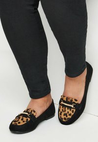 Mustat mokkasiinit, joissa on leopardikuosiset mokkatossut, ja kultasävyinen koriste kengän päällä. Litteä pohja, pyöreä kärki.