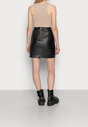 Frau trägt ein beiges ärmelloses Top, einen schwarzen Leder-Minirock mit Reißverschluss hinten und schwarze klobige Stiefeletten, steht vor einer weißen Wand.