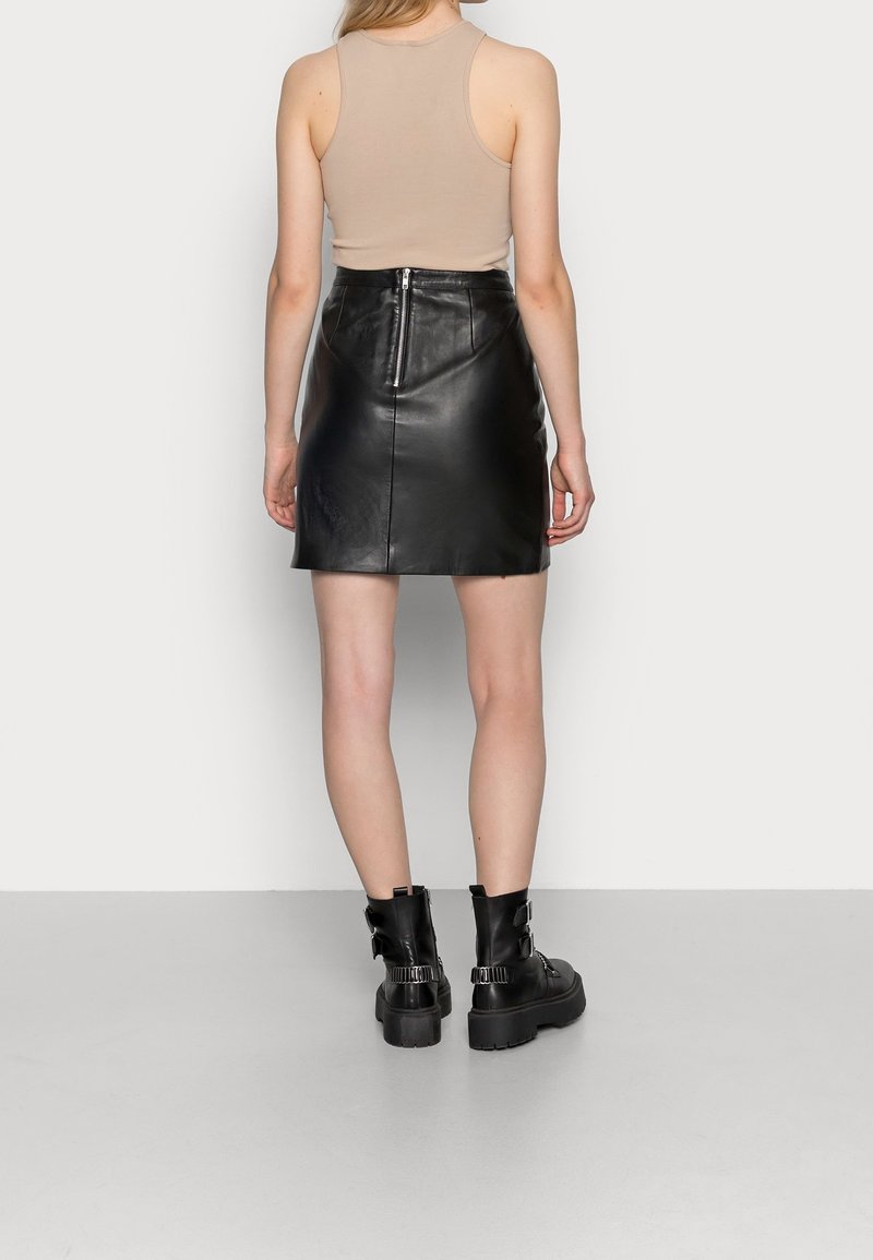 Femme portant un haut beige sans manches, une mini-jupe en cuir noire avec fermeture éclair au dos, et des bottines noires épaisses, debout contre un mur blanc.
