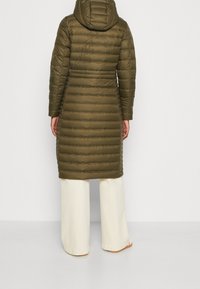 Persoon in een olijfgroene lange gewatteerde jas met capuchon en witte wijde broek, staand met de rug naar de kijker op een effen lichte achtergrond.