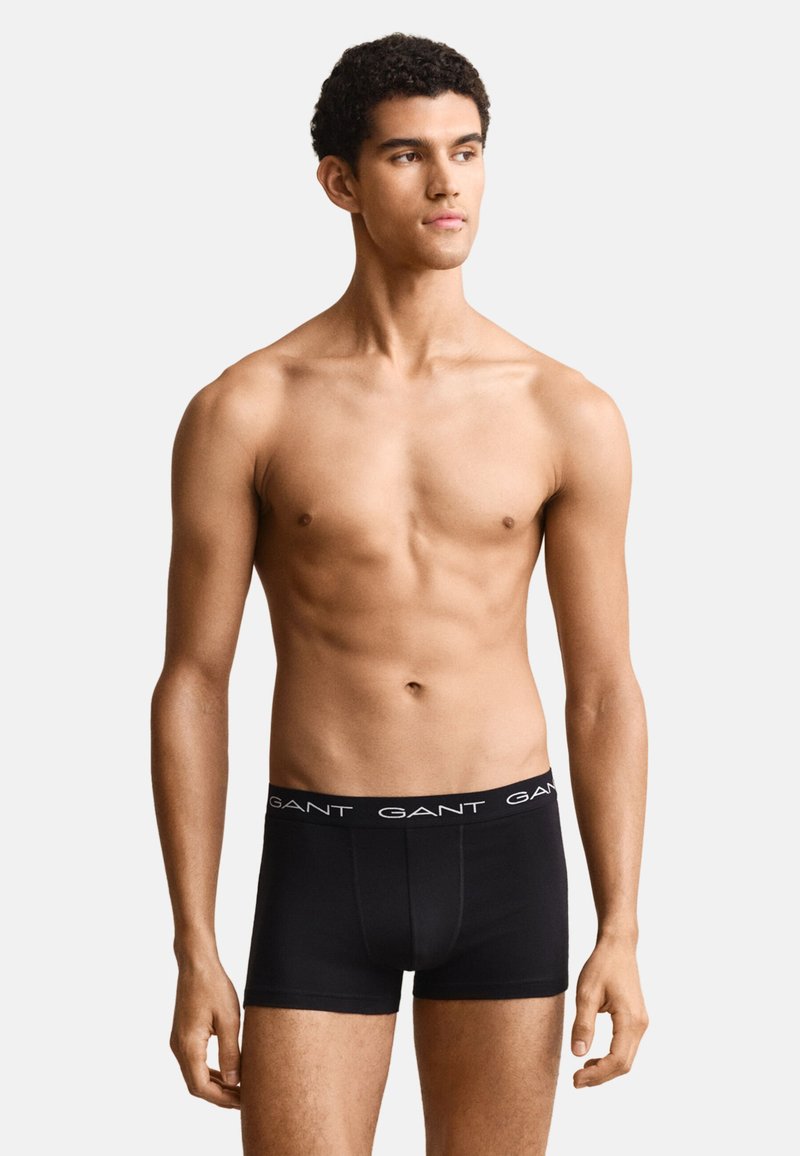 Junger Mann mit kurzen lockigen Haaren, der schwarze GANT Boxershorts trägt, steht und schaut leicht nach rechts vor einem schlichten Hintergrund.