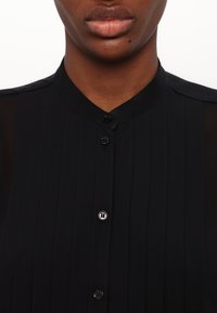 Camicia nera plissettata con colletto arrotondato, chiusura frontale con bottoni neri e dettagli in tessuto trasparente sulle spalle.