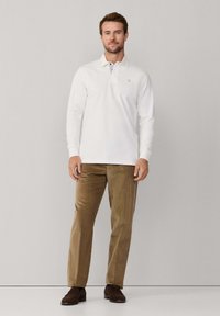 Witte lange mouw polo shirt met een kraag, gecombineerd met bruine ribfluweel broeken. De outfit heeft een ontspannen pasvorm en wordt aangevuld met bruine suede instappers.