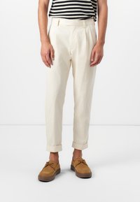 Pantalones plisados de color crema con puños enrollados, hechos de una tela ligera. Llevados con zapatos de ante marrones, combinados con una blusa a rayas.