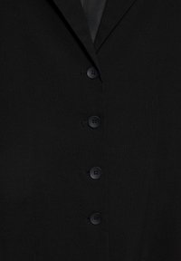Veste de costume noire avec quatre boutons noirs fermés, laissant apparaître une partie d'une chemise noire et d'une cravate noire en dessous.