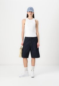 Vit linne, mörka denim-kortbyxor som når knäna, ljusblå keps, vita sneakers och krämfärgad väska. Modellen står mot en neutral bakgrund.