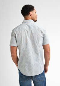 La camicia a maniche corte in tessuto grigio chiaro presenta un motivo a pois blu. Il retro ha un colletto classico e un orlo curvo.