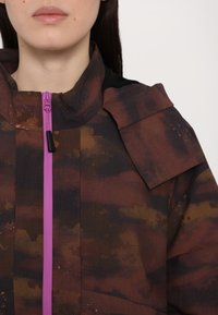 Wasserabweisende Jacke in dunkelbraun mit rostfarbenen Mustern, ausgestattet mit einem lila Reißverschluss, hohem Kragen und einem einzigartigen Schulterklappen-Design.