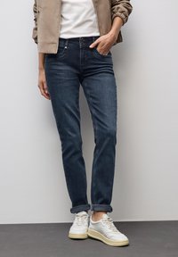 Henkilö, joka wear nousevat tummansiniset farkut, valkoiset tennarit, beige takki ja valkoinen paita, seisoo tasaista seinää vasten.