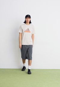 Tricou Adidas bej deschis cu logo portocaliu, pantaloni scurți gri, șosete albe și pantofi negri. Culori solide, croială casual, design minim.