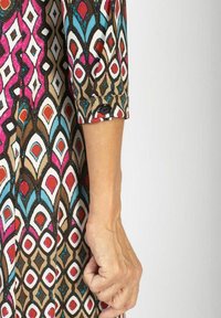 Wielokolorowa sukienka w wzory z brązowym tłem, z geometrycznymi wzorami w różowym, niebieskim, czerwonym i białym; długie rękawy z detalami w postaci guzików.