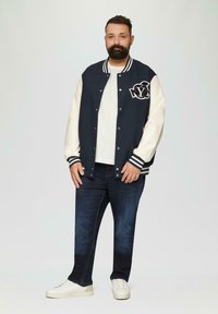 Marineblauwe en crèmekleurige varsityjack met gestreepte ribbing, voorzien van een geborduurd NY-logo, gecombineerd met donkerdenim jeans en witte sneakers.