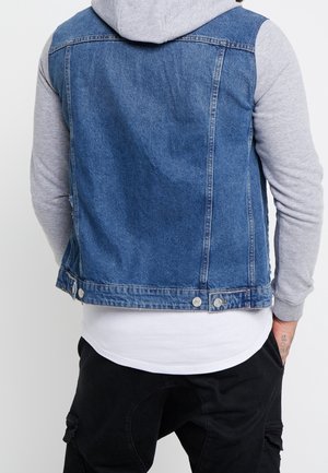 Persona con chaleco vaquero azul sobre una sudadera con capucha gris, camisa blanca debajo y pantalones negros, vista desde atrás con las manos en los bolsillos.