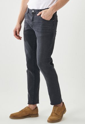 Homme portant un jean gris foncé slim et des chaussures à lacets en daim marron clair, avec une main dans la poche, debout devant un fond uni.