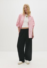 Mladá žena s blond vlasmi má na sebe ružový pruhovaný tričko cez biele crop top, čierne široké nohavice, biele tenisky a vrstvené náhrdelníky.