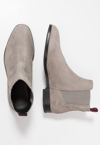 Une paire de bottes Chelsea en daim gris avec des panneaux latéraux élastiques et des semelles noires, une botte vue de dessus, l'autre de côté.