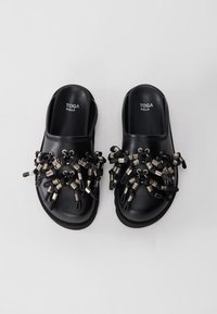 Black leather slide sandals featuring an intricate arrangement of black cords and metallic accents on the upper. Smooth texture, flat sole.
