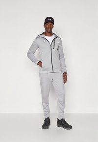 Casaco cinzento claro com fecho éclair, detalhes pretos e logótipo, combinado com calças a condizer. Veste uma t-shirt branca por baixo e ténis pretos.