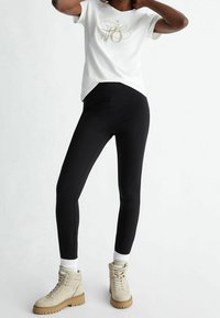 Vit t-shirt med guldgrafik, kombinerad med svarta leggings och beige ankelboots med strukturerad sula och snörningsdetalj.