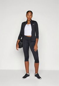 Sort atletikjakke med lynlås, hvid crop top, sorte capri-leggings og sorte sneakers. Glat stof og figursyet design.