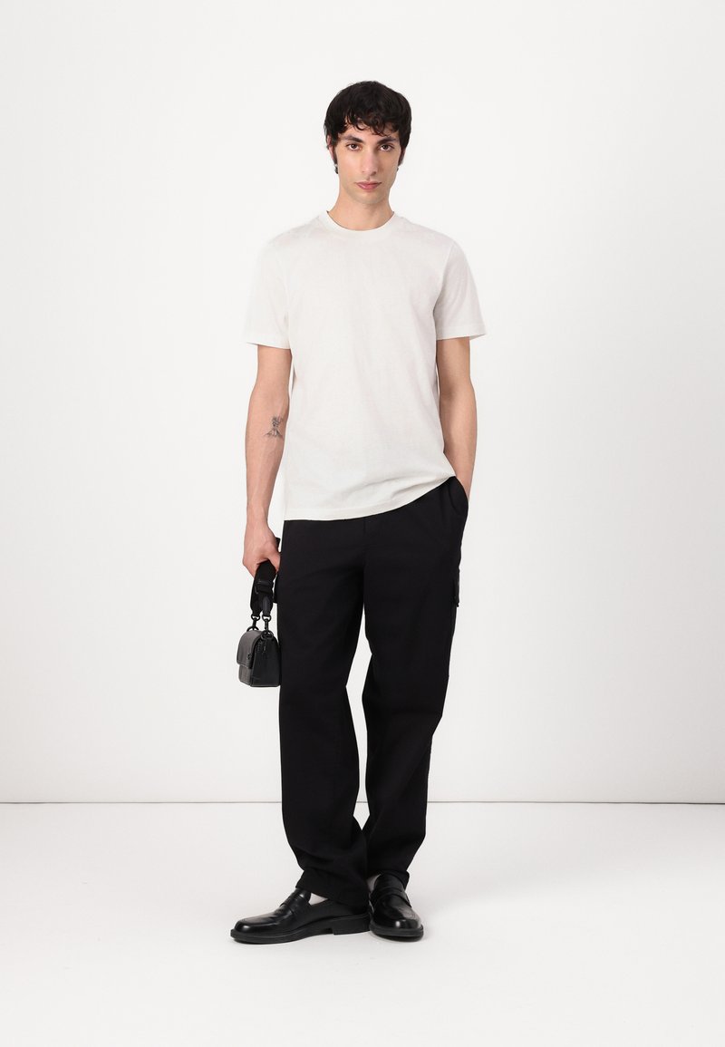 Weißes T-Shirt mit kurzen Ärmeln, schwarze weit geschnittene Hose, schwarze Schuhe, hält eine kleine schwarze Tasche. Glatter Stoff, minimalistisches Design, neutrale Farben.
