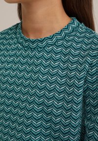 Tyrkysový a bílý vzorovaný top s designem ševron, kulatým výstřihem a texturovaným pleteným materiálem. Pohled na detaily oděvu.