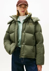 Doudoune vert olive avec une coupe ample, des boutons-pression et un col montant. Portée par-dessus une chemise rayée. Tissu durable avec une texture douce.