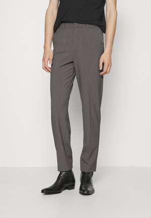 Mężczyzna noszący wąskie, szare eleganckie spodnie, czarne buty Chelsea i czarną koszulę, stojący na białym tle.