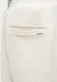 Κοντινή λήψη πίσω τσέπης σε παντελόνι εκρού χρώματος με μια μικρή λευκή ετικέτα που γράφει "JACK & JONES" ραμμένη κοντά στην άκρη της τσέπης.