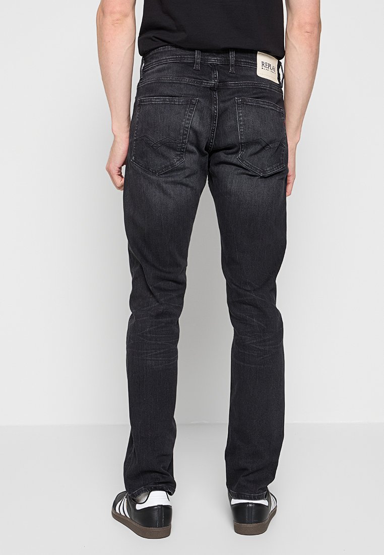 Man in slim-fit zwarte jeans en zwarte sneakers met witte strepen, staand tegen een effen lichtgrijze achtergrond.