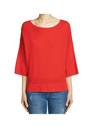Rotes Strickoberteil mit weiter Bootsausschnitt, Dreiviertelärmeln und einem strukturierten Muster. Kombiniert mit blauen Jeans.