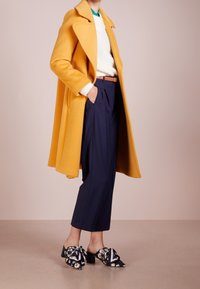 Cappotto di lana giallo, maglione a maglia bianco, pantaloni plissettati blu navy e mule floreali con base nera e dettaglio a fiocco, su uno sfondo neutro.