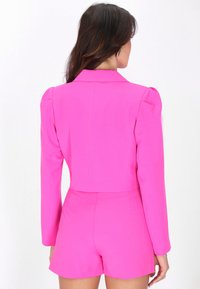 Frau mit langen dunklen Haaren, die einen leuchtend pinken Blazer und passende hoch taillierte Shorts trägt, von hinten vor einem weißen Hintergrund zu sehen.