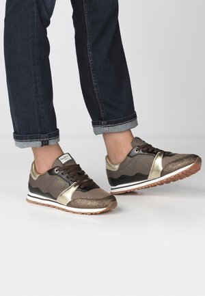 Osoba ubrana w podwinięte ciemnoniebieskie dżinsy oraz brązowo-złote trampki sznurowane na białej podłodze.