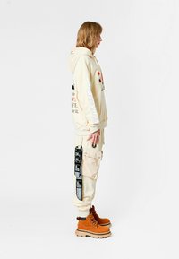 Persona con sudadera beige y pantalones cargo con parches negros, de pie de lado, usando botas de cordones color beige, contra un fondo blanco.