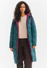 Azurově modrý prošívaný dlouhý kabát s vínovou podšívkou, vybavený kapucí a předním zipem. Nošený přes béžový svetr s copánkovým vzorem a tmavé kalhoty.