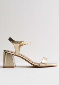 Sandalia de tacón metálica dorada con tacón cuadrado, correa ajustable en el tobillo y dos bandas frontales delgadas. Textura suave y diseño minimalista.
