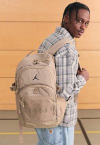 Mochila bege com vários bolsos com fecho, detalhes em rede e um logótipo preto. Tecido durável com alças ajustáveis. Usada num ombro.