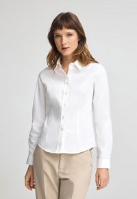 Weißes Hemd mit langen Ärmeln, taillierte Passform und einem subtilen gestickten Logo auf der linken Brust. Glatter Stoff und umgeschlagene Handgelenke.