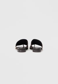 Μαύρα επίπεδα σανδάλια με δαχτυλίδι δαχτύλου και χρυσές διακοσμητικές πινελιές με καρφιά, εμφανιζόμενα από πίσω σε απλό λευκό φόντο.