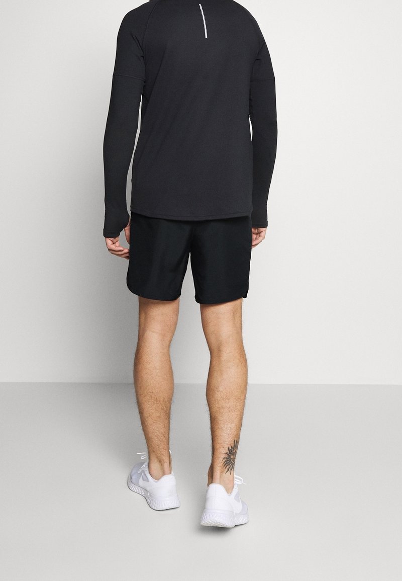 Homme portant un haut de sport noir à manches longues, un short noir, des baskets blanches, debout dos à la caméra montrant un tatouage à la cheville.
