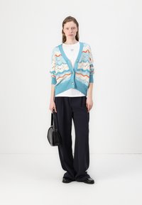 Strikket cardigan i lyseblå, kremfarge og orange med bølge mønstre. Har en løs passform, V-hals og knappestengning. Kombinert med mørke bukser.