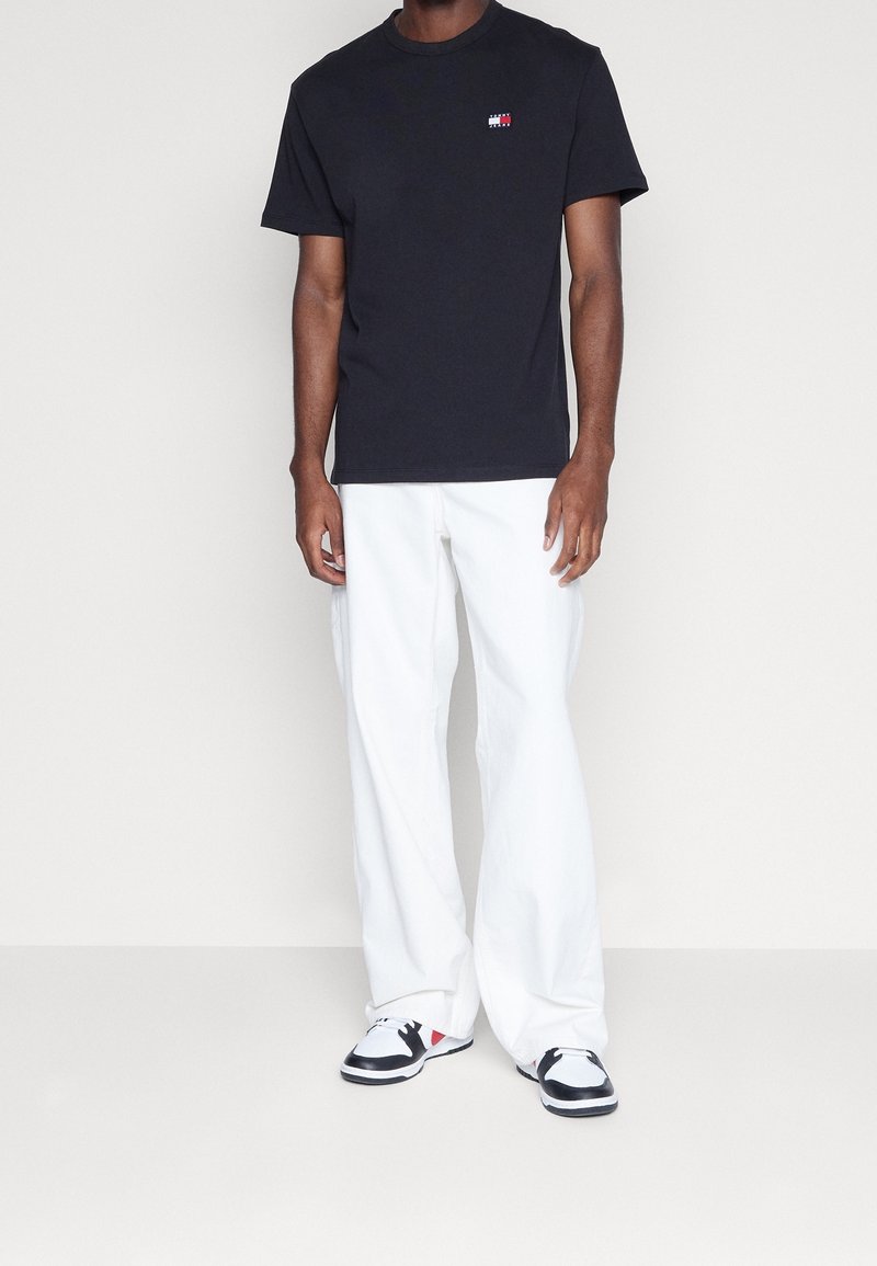 Czarna bawełniana koszulka z okrągłym dekoltem, z niewielkim wielokolorowym logo. W połączeniu z białymi szerokimi spodniami i czarnymi tenisówkami.