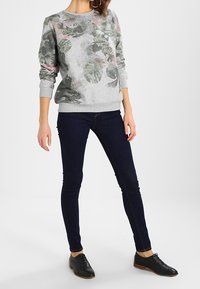 Kobieta ubrana w szary sweter z zielonymi i różowymi wzorami liści, ciemne skinny jeansy i czarne buty sznurowane, stojąca na tle białym.