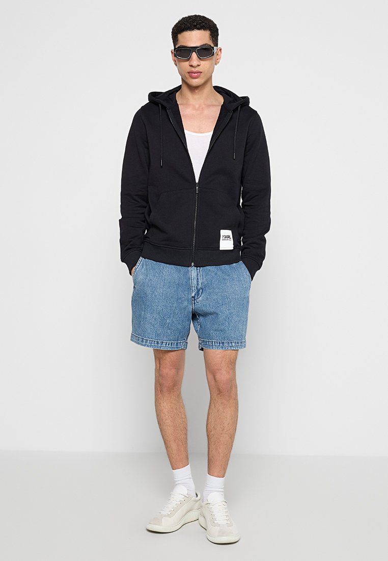 Felpa nera con zip, dotata di tasca frontale e etichetta con logo, abbinata a pantaloni corti in denim azzurro e sneakers bianche. Il modello indossa occhiali da sole.